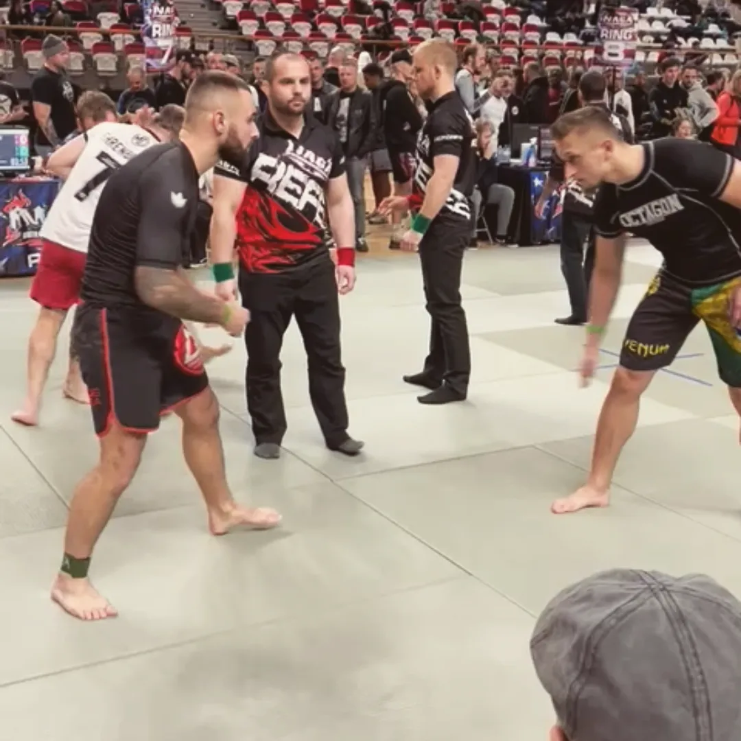 Twee BJJ-deelnemers staan klaar voor hun partij op de wedstrijdmat, met de scheidsrechter tussen hen.